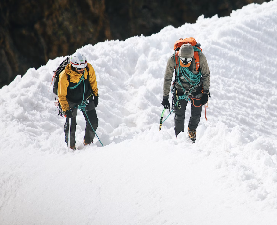 Two climbers equipped with ice axes and ropes ascending a snowy mountain slope, dressed in warm protective gear, including helmets and backpacks, against a snowy alpine backdrop.